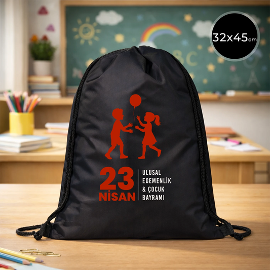 32x45 cm siyah büzgülü sırt çantası üzerinde kırmızı “23 Nisan Ulusal Egemenlik ve Çocuk Bayramı” baskısı, balonlu çocuk figürleri ve sınıf temalı arka plan