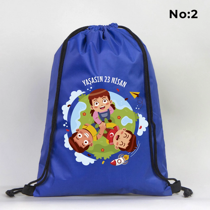 32x45 cm mavi büzgülü sırt çantası üzerinde dünya figürü, renkli çocuk silüetleri ve “23 Nisan Ulusal Egemenlik ve Çocuk Bayramı Kutlu Olsun” yazılı baskı, sınıf temalı arka plan