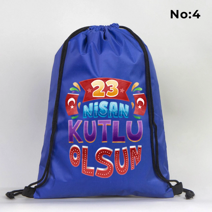 32x45 cm mavi büzgülü sırt çantası üzerinde dünya figürü, renkli çocuk silüetleri ve “23 Nisan Ulusal Egemenlik ve Çocuk Bayramı Kutlu Olsun” yazılı baskı, sınıf temalı arka plan