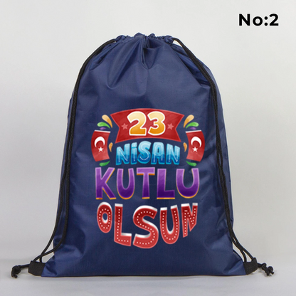 32x45 cm lacivert büzgülü sırt çantası üzerinde “Yaşasın 23 Nisan” yazılı çocuk temalı renkli baskı, roket ve kâğıt uçak detayları, kutlama temalı sınıf arka planı
