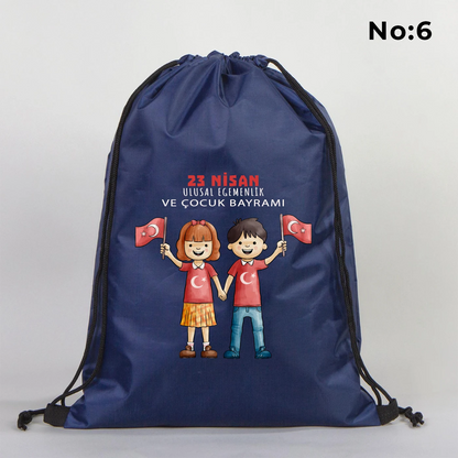 32x45 cm lacivert büzgülü sırt çantası üzerinde “Yaşasın 23 Nisan” yazılı çocuk temalı renkli baskı, roket ve kâğıt uçak detayları, kutlama temalı sınıf arka planı