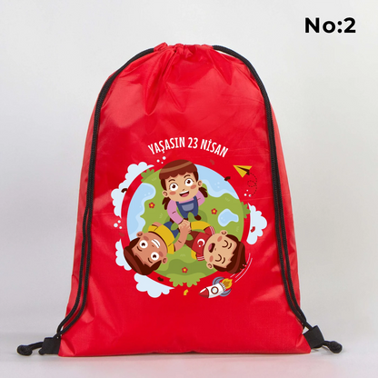 32x45 cm kırmızı büzgülü sırt çantası üzerinde beyaz “23 Nisan Ulusal Egemenlik ve Çocuk Bayramı” baskısı, balonlu çocuk figürleri ve kutlama temalı sınıf arka planı