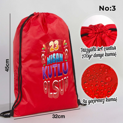 32x45 cm kırmızı büzgülü sırt çantası üzerinde beyaz “23 Nisan Ulusal Egemenlik ve Çocuk Bayramı” baskısı, balonlu çocuk figürleri ve kutlama temalı sınıf arka planı