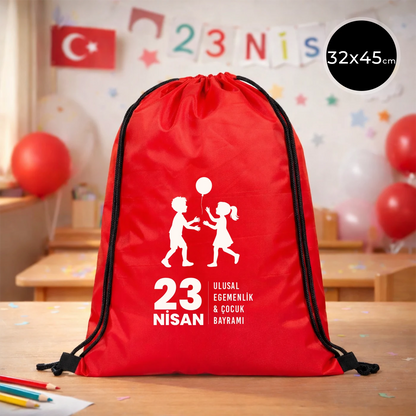 32x45 cm kırmızı büzgülü sırt çantası üzerinde beyaz “23 Nisan Ulusal Egemenlik ve Çocuk Bayramı” baskısı, balonlu çocuk figürleri ve kutlama temalı sınıf arka planı