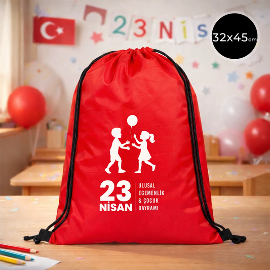32x45 cm kırmızı büzgülü sırt çantası üzerinde beyaz “23 Nisan Ulusal Egemenlik ve Çocuk Bayramı” baskısı, balonlu çocuk figürleri ve kutlama temalı sınıf arka planı
