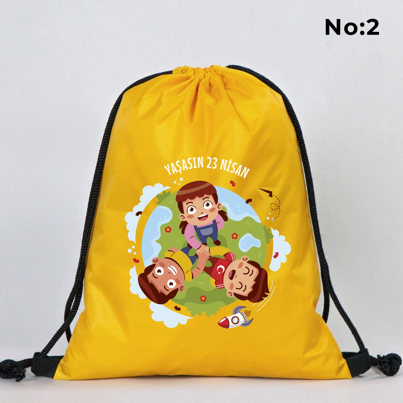 32x45 cm sarı büzgülü sırt çantası üzerinde “23 Nisan Kutlu Olsun” yazılı renkli çocuk figürleri baskısı, balon detayları ve kutlama temalı sınıf arka planı