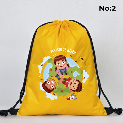 32x45 cm sarı büzgülü sırt çantası üzerinde “23 Nisan Kutlu Olsun” yazılı renkli çocuk figürleri baskısı, balon detayları ve kutlama temalı sınıf arka planı