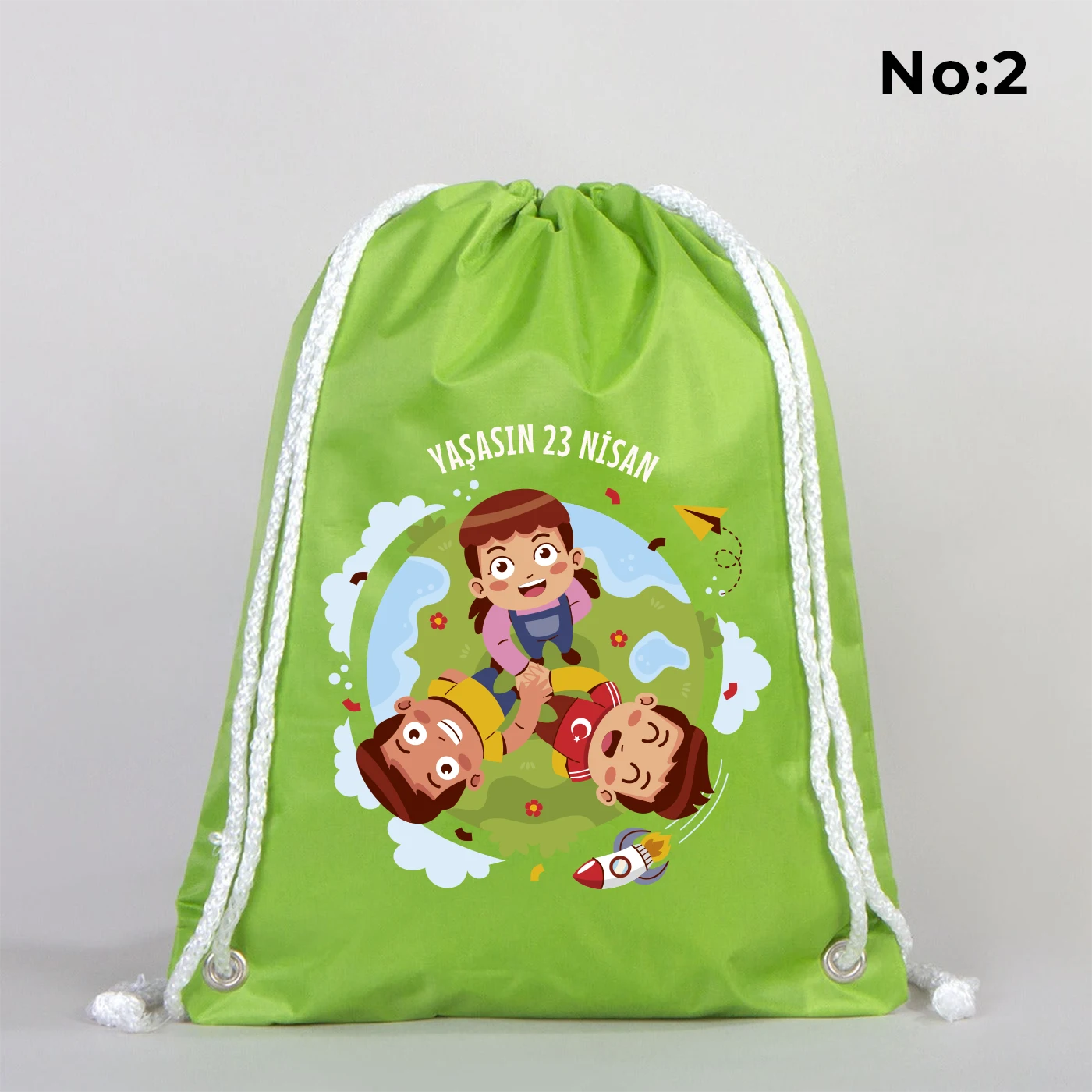 32x45 cm yeşil büzgülü sırt çantası üzerinde dünya figürü, renkli çocuk silüetleri ve “23 Nisan Ulusal Egemenlik ve Çocuk Bayramı Kutlu Olsun” yazılı baskı, kutlama temalı sınıf arka planı
