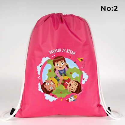 32x45 cm pembe büzgülü sırt çantası üzerinde “23 Nisan Kutlu Olsun” yazılı renkli çocuk figürleri baskısı, balon detayları ve kutlama temalı sınıf arka planı