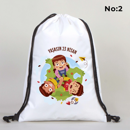 32x45 cm beyaz büzgülü sırt çantası üzerinde kırmızı “23 Nisan Ulusal Egemenlik ve Çocuk Bayramı” baskısı, balonlu çocuk figürleri ve kutlama temalı sınıf arka planı