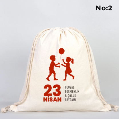 35x40 cm naturel renkli büzgülü bez çanta üzerinde “Yaşasın 23 Nisan” yazılı çocuk temalı renkli baskı, roket ve kâğıt uçak detayları, 23 Nisan süslemeli sınıf arka planı