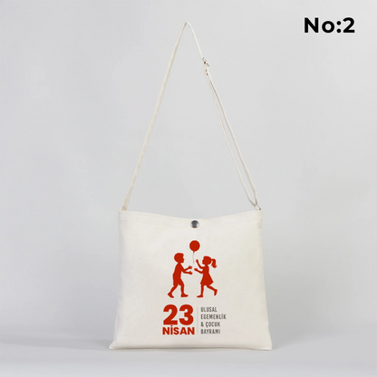 35x30 cm krem renkli çapraz askılı kumaş çanta üzerinde çıtçıt kapama detayı, “23 Nisan Kutlu Olsun” yazılı renkli çocuk figürleri baskısı ve sınıf temalı arka plan