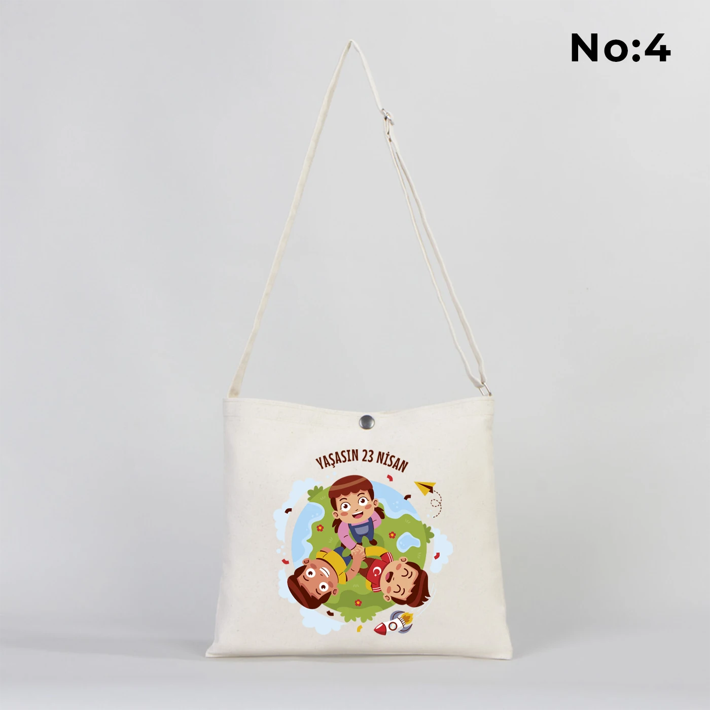 35x30 cm krem renkli çapraz askılı kumaş çanta üzerinde çıtçıt kapama detayı, “23 Nisan Kutlu Olsun” yazılı renkli çocuk figürleri baskısı ve sınıf temalı arka plan