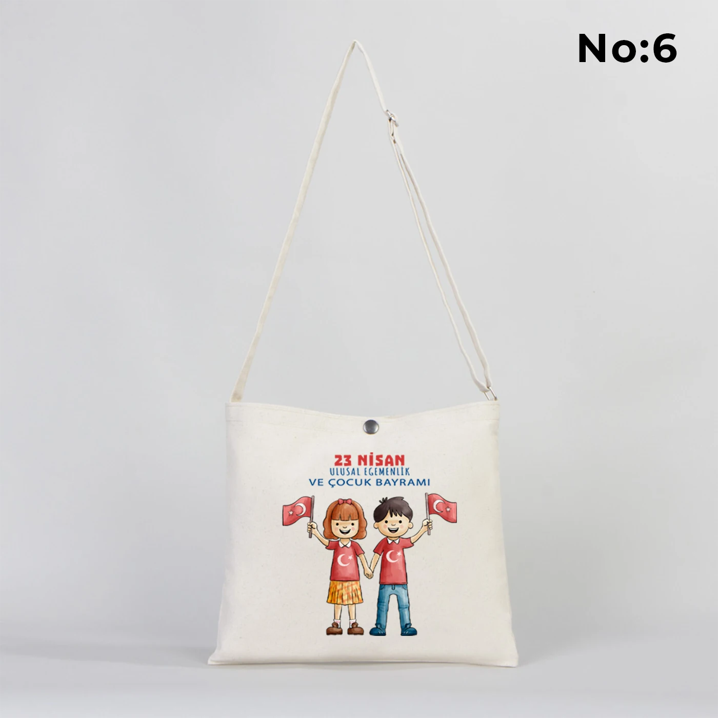 35x30 cm krem renkli çapraz askılı kumaş çanta üzerinde çıtçıt kapama detayı, “23 Nisan Kutlu Olsun” yazılı renkli çocuk figürleri baskısı ve sınıf temalı arka plan