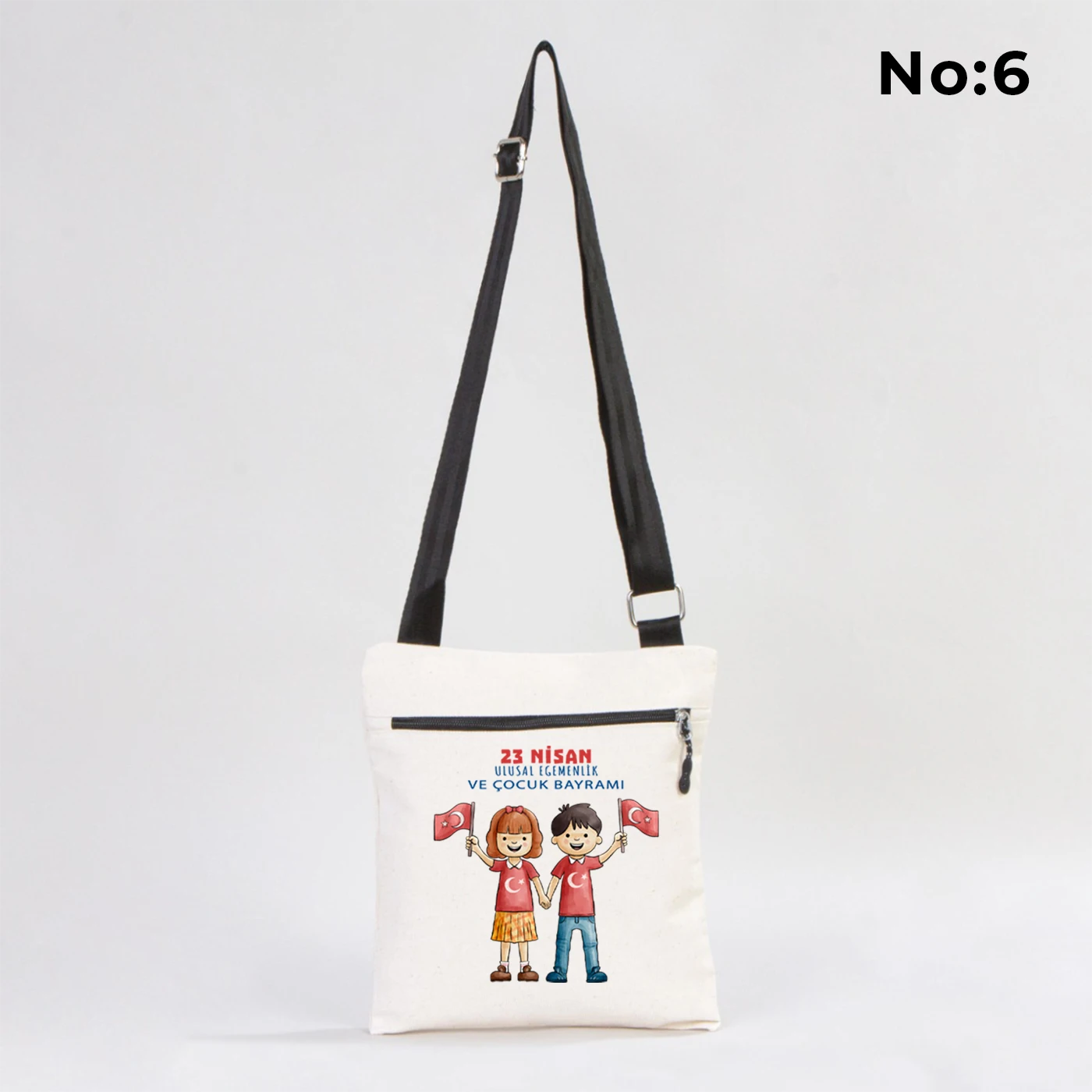 25x28 cm krem renkli çapraz askılı kumaş çanta üzerinde fermuarlı ön cep, “Yaşasın 23 Nisan” yazılı çocuk temalı renkli baskı ve sınıf ortamı arka planı