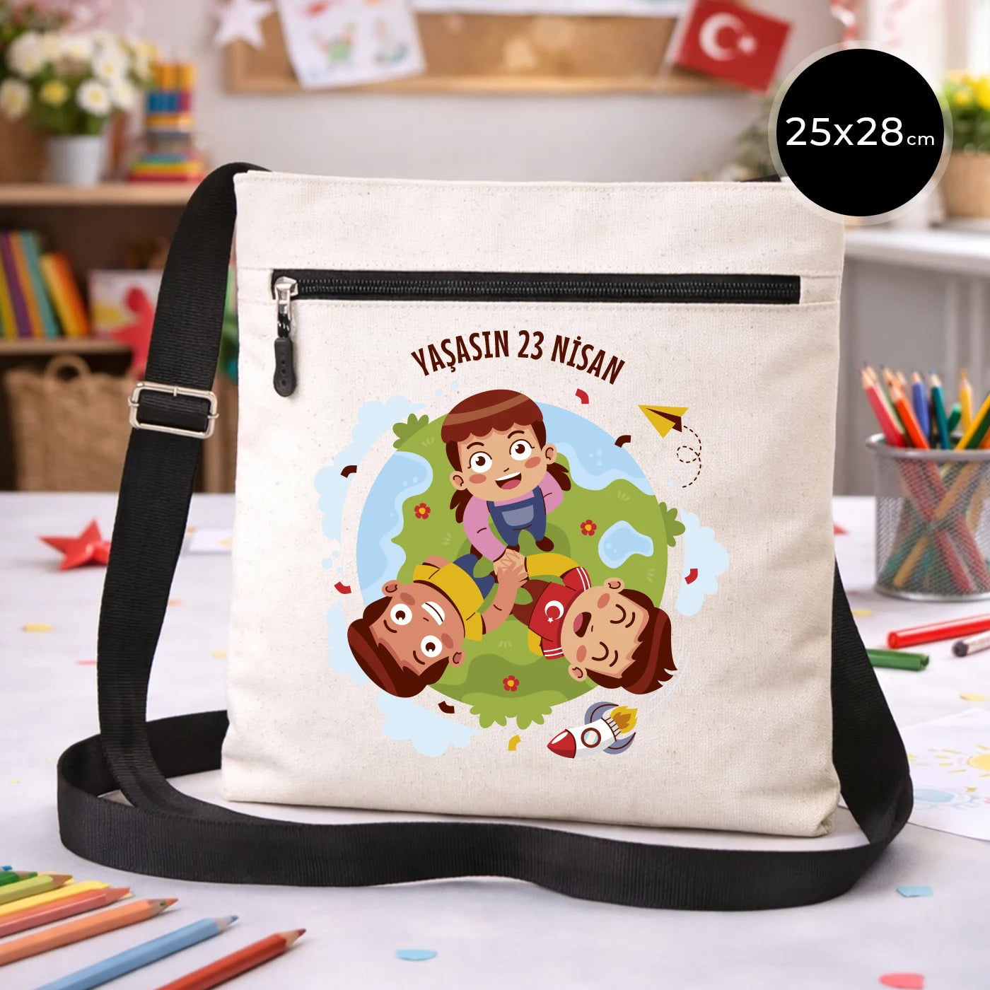 25x28 cm krem renkli çapraz askılı kumaş çanta üzerinde fermuarlı ön cep, “Yaşasın 23 Nisan” yazılı çocuk temalı renkli baskı ve sınıf ortamı arka planı