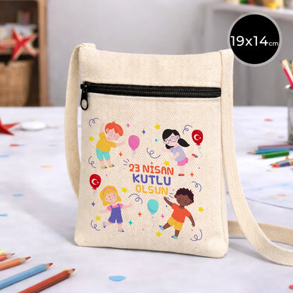 19x14 cm krem renkli çapraz askılı kumaş çanta üzerinde “23 Nisan Kutlu Olsun” yazısı, balonlu çocuk figürleri baskısı ve fermuarlı ön cep detayı