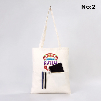 35x40 cm ön cepli ham bez çanta üzerinde “Yaşasın 23 Nisan” yazılı çocuk illüstrasyonu; ön cepte renkli kalemler, küçük Türk bayrağı ve rulo kâğıt bulunan 23 Nisan temalı bez çanta.