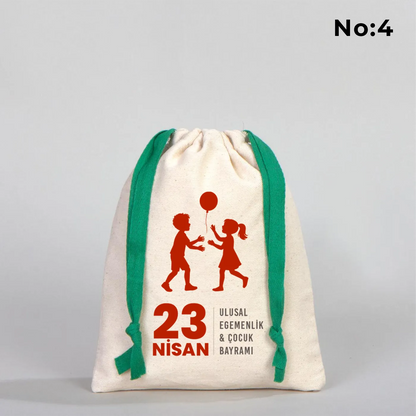 Naturel renkli, yeşil büzgü ipli bez kese üzerinde “Yaşasın 23 Nisan” yazılı çocuk temalı renkli baskı, roket ve kâğıt uçak detayları
