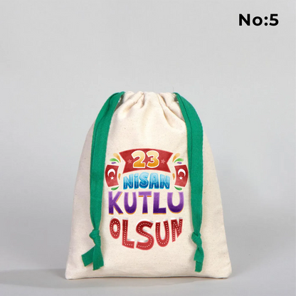 Naturel renkli, yeşil büzgü ipli bez kese üzerinde “Yaşasın 23 Nisan” yazılı çocuk temalı renkli baskı, roket ve kâğıt uçak detayları