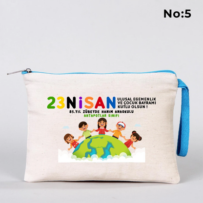 23 Nisan temalı 21x15 cm fermuarlı bez kalemlik üzerinde “23 Nisan Ulusal Egemenlik ve Çocuk Bayramı Kutlu Olsun” yazısı, ay ve yıldız üzerinde oturan iki çocuk illüstrasyonu ve okul - sınıf özelleştirilebilir alan.