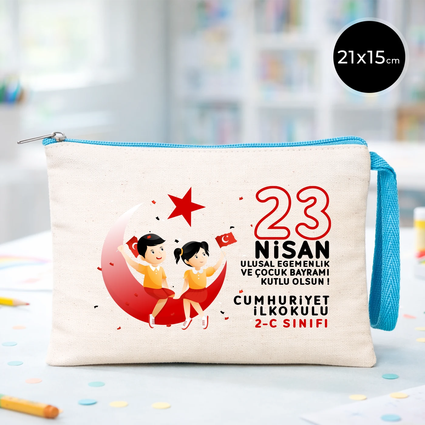 23 Nisan temalı 21x15 cm fermuarlı bez kalemlik üzerinde “23 Nisan Ulusal Egemenlik ve Çocuk Bayramı Kutlu Olsun” yazısı, ay ve yıldız üzerinde oturan iki çocuk illüstrasyonu ve okul - sınıf özelleştirilebilir alan.