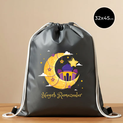 32x45 cm antrasit imperteks büzgülü sırt çantası, ip büzgülü/omuz askılı model; ön yüzünde “Hayırlı Ramazanlar” yazısı ve hilal–cami temalı renkli baskı bulunan Bayram Çantam hediyelik çanta.