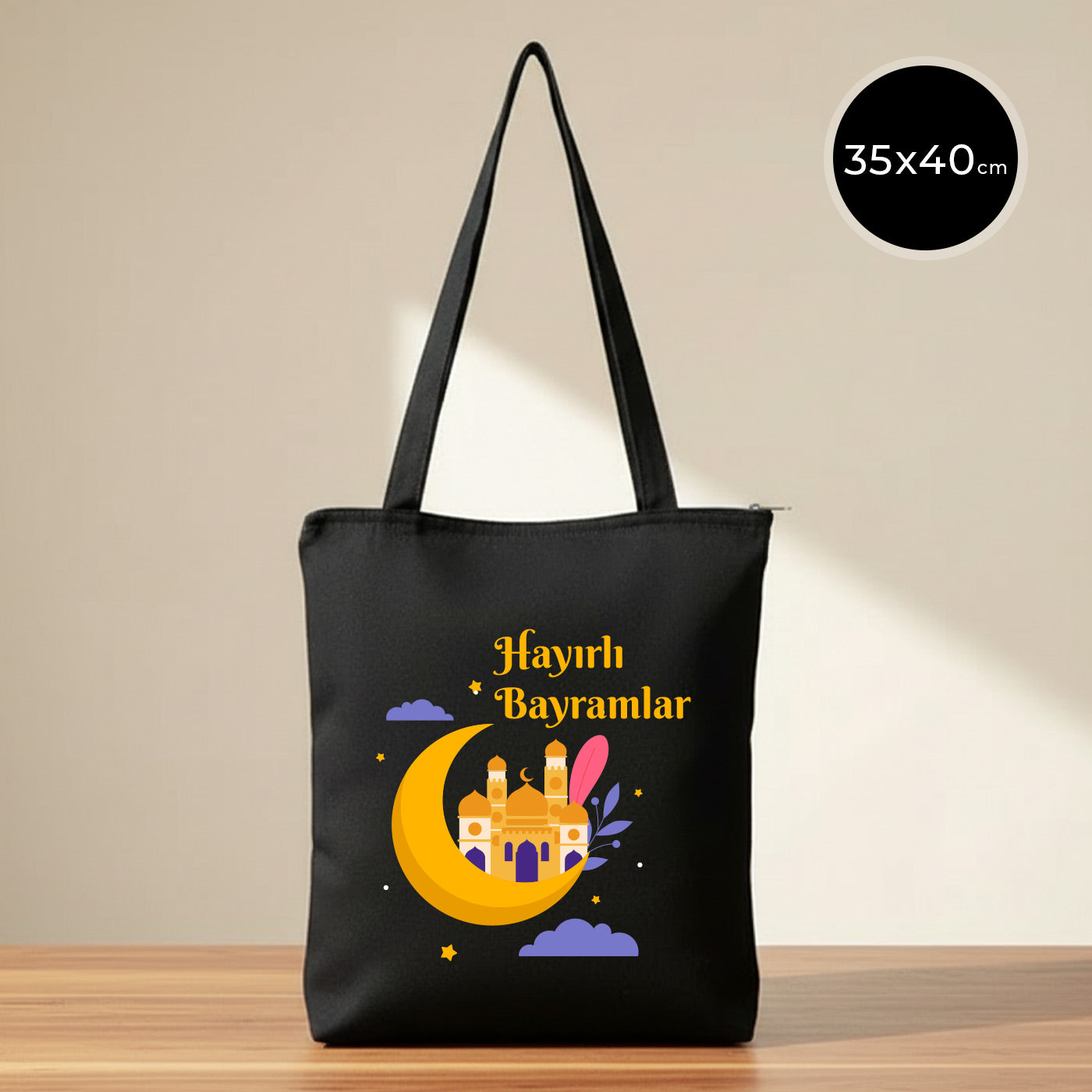 35x40 cm siyah gabardin fermuarlı tote çanta; ön yüzde altın sarısı hilal üzerinde cami silüeti, mor bulut detayları ve “Hayırlı Bayramlar” yazısı baskısı, omuz askılı günlük bez çanta.