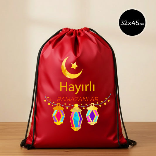 32×45 cm ölçülerinde kırmızı ip büzgülü bez sırt çantası üzerinde üstte sarı hilal ve yıldız, altta üç adet renkli asılı fener illüstrasyonu ve ortada “Hayırlı Ramazanlar” yazısı; siyah ip askılar ve sade açık renkli arka plan.