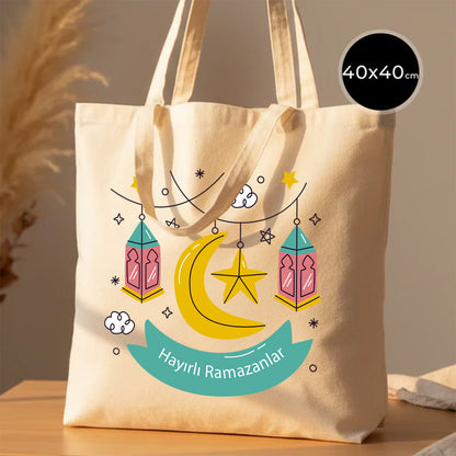 40×40 cm ölçülerinde doğal ham bez tote çanta üzerinde asılı pembe–turkuaz fenerler, sarı hilal ve yıldız illüstrasyonu ile turkuaz şerit içinde “Hayırlı Ramazanlar” yazısı; arka planda ahşap yüzey ve sıcak ışık.