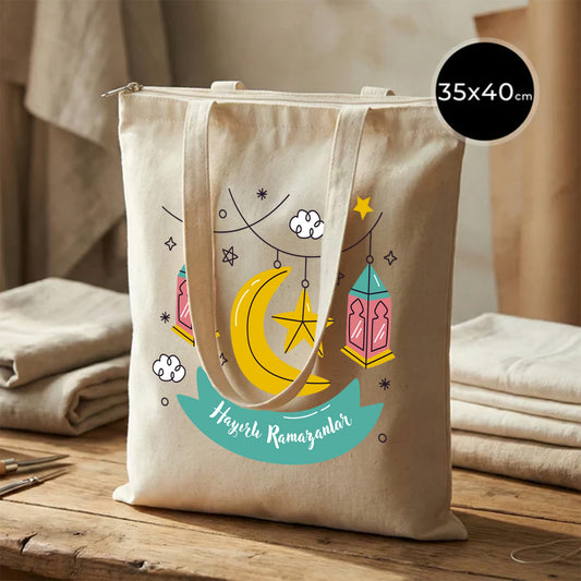 35×40 cm ölçülerinde doğal ham bez fermuarlı tote çanta üzerinde asılı renkli fenerler, yıldızlar ve hilal ile birlikte turkuaz şerit içinde “Hayırlı Ramazanlar” yazısı; ahşap zemin üzerinde katlı kumaşların yanında.