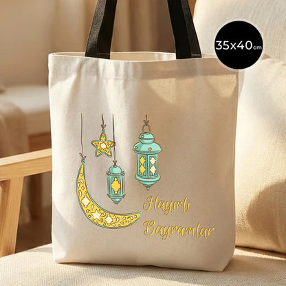 35×40 cm ölçülerinde doğal ham bez tote çanta üzerinde asılı turkuaz fenerler, sarı yıldız ve hilal illüstrasyonu ile sağ altında “Hayırlı Bayramlar” yazısı; arka planda ahşap sandalye ve açık renkli iç mekân.