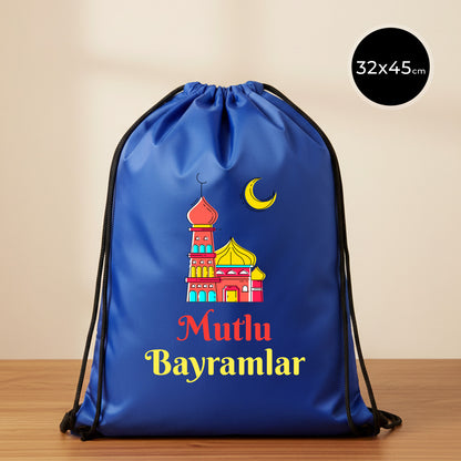 32×45 cm ölçülerinde mavi ip büzgülü bez sırt çantası üzerinde kırmızı–sarı tonlarında cami illüstrasyonu, sarı hilal ve altında “Mutlu Bayramlar” yazısı; siyah ip askılar ve sade açık renkli arka plan.