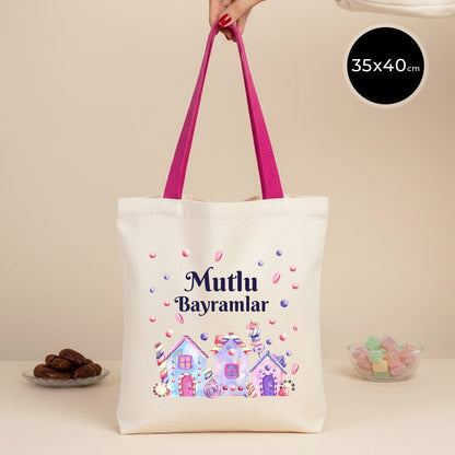 35×40 cm ölçülerinde doğal ham bez tote çanta, fuşya renkli askıları bir el tarafından tutuluyor; çantanın ön yüzünde pastel renkli şeker evleri, şeker taneleri ve üstte “Mutlu Bayramlar” yazısı, yanında renkli lokum kâsesi.