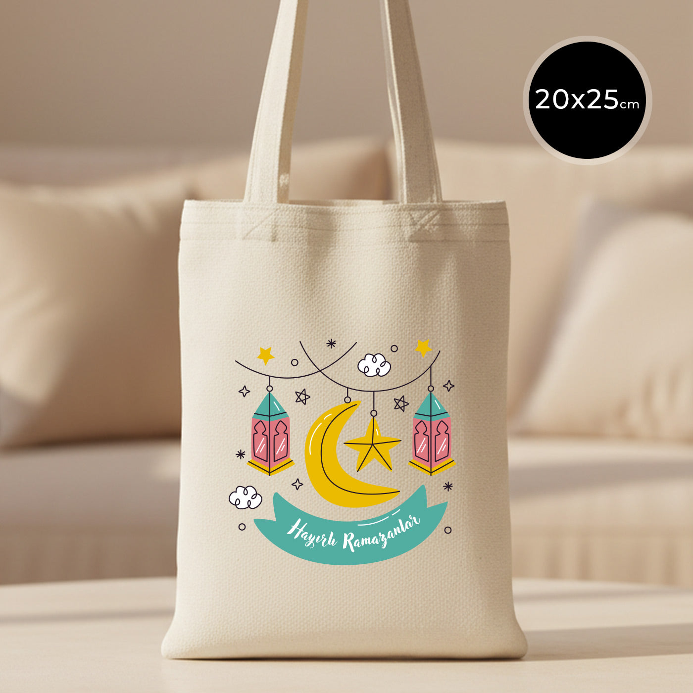 20×25 cm ölçülerinde doğal ham bez tote çanta üzerinde asılı fenerler, yıldızlar ve hilal ile birlikte turkuaz şerit içinde “Hayırlı Ramazanlar” yazısı; arka planda açık renkli kanepe.