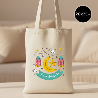 20×25 cm ölçülerinde doğal ham bez tote çanta üzerinde asılı fenerler, yıldızlar ve hilal ile birlikte turkuaz şerit içinde “Hayırlı Ramazanlar” yazısı; arka planda açık renkli kanepe.
