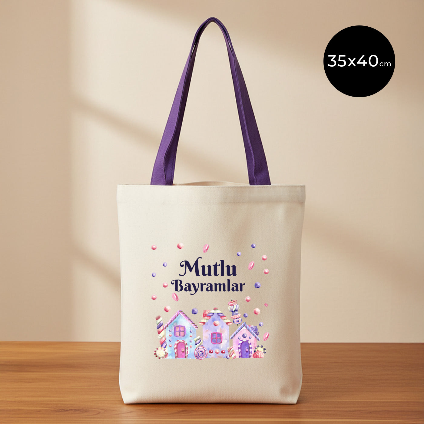 35×40 cm ölçülerinde doğal ham bez tote çanta üzerinde mor askılar; ön yüzünde pastel renkli şeker evleri, etrafa saçılmış şeker taneleri ve üstte “Mutlu Bayramlar” yazısı, sade açık renkli arka plan ve ahşap yüzey.