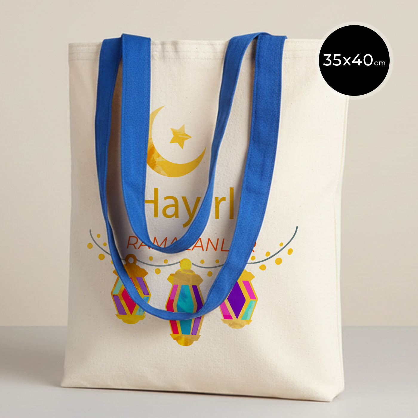 35×40 cm ölçülerinde doğal ham bez tote çanta üzerinde mavi askılar; ön yüzünde sarı hilal ve yıldız ile altında üç adet renkli asılı fener illüstrasyonu ve ortada “Hayırlı Ramazanlar” yazısı, sade açık renkli arka plan.