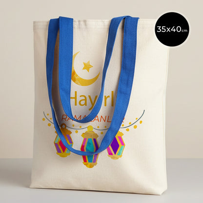 35×40 cm ölçülerinde doğal ham bez tote çanta üzerinde mavi askılar; ön yüzünde sarı hilal ve yıldız ile altında üç adet renkli asılı fener illüstrasyonu ve ortada “Hayırlı Ramazanlar” yazısı, sade açık renkli arka plan.