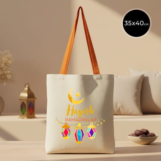 35×40 cm ölçülerinde doğal ham bez tote çanta üzerinde sarı hilal ve yıldız ile alt kısımda renkli asılı fener illüstrasyonları ve “Hayırlı Ramazanlar” yazısı; arka planda açık renkli kanepe ve yanında hurma kâsesi.