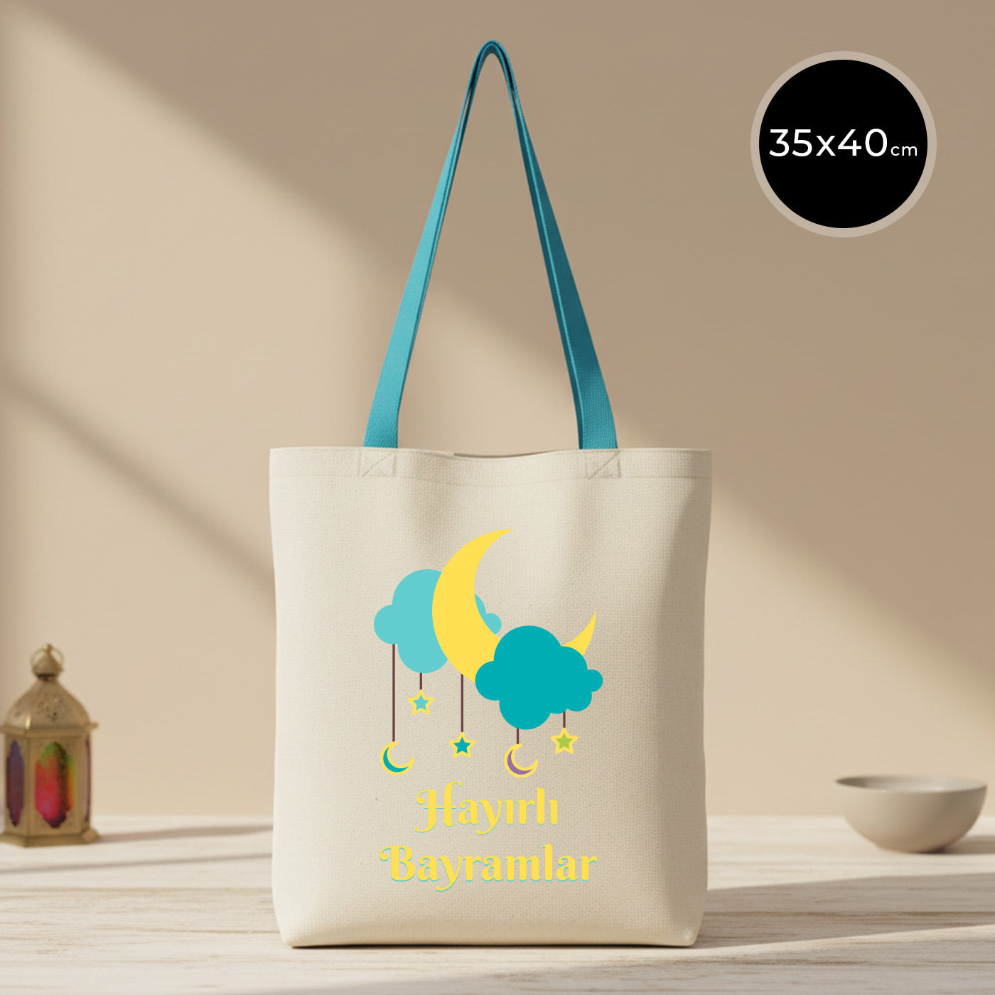 35×40 cm ölçülerinde doğal ham bez tote çanta üzerinde turkuaz bulutlar, sarı hilal ve sarkık yıldız illüstrasyonlarıyla “Hayırlı Bayramlar” yazısı; turkuaz askılı, arka planda sade açık renkli masa ve kâse.