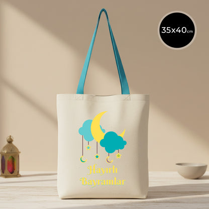 35×40 cm ölçülerinde doğal ham bez tote çanta üzerinde turkuaz bulutlar, sarı hilal ve sarkık yıldız illüstrasyonlarıyla “Hayırlı Bayramlar” yazısı; turkuaz askılı, arka planda sade açık renkli masa ve kâse.