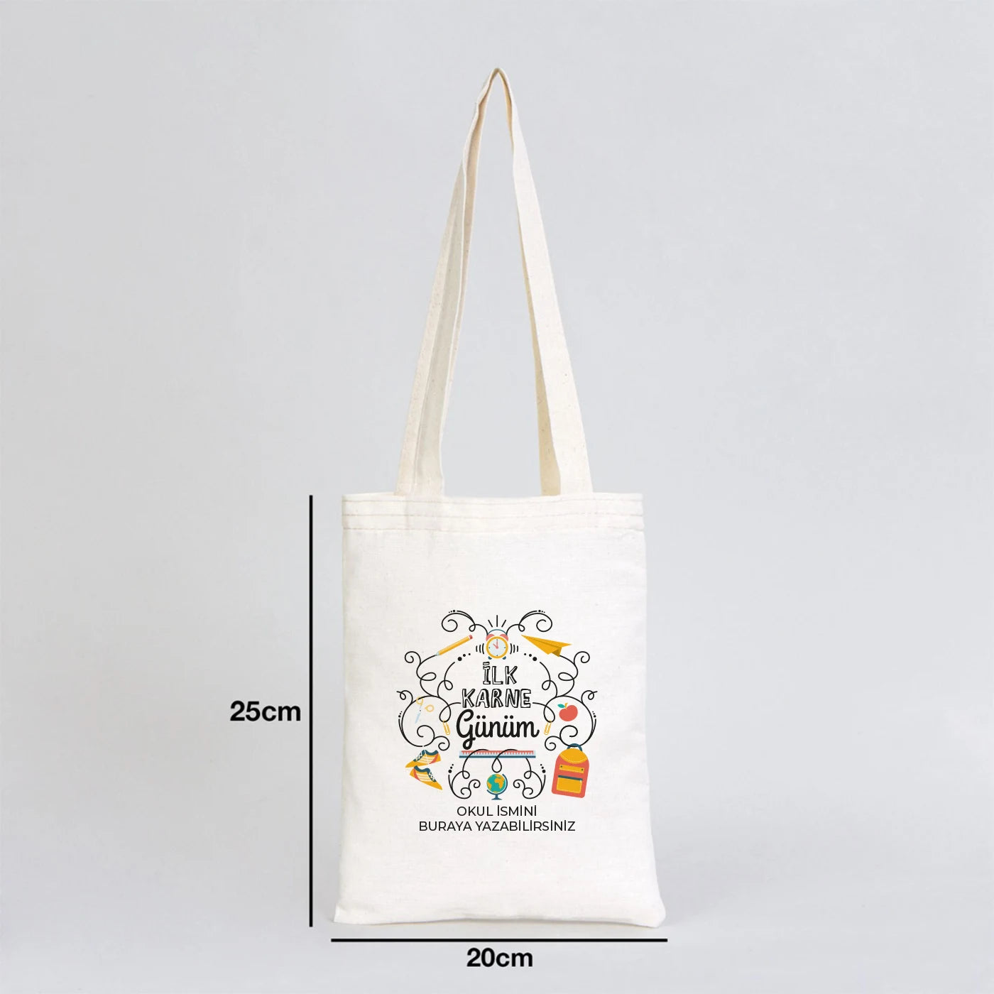 Sıcak ve renkli bir sınıf ortamında, ahşap masada sergilenen 20x25 cm ham pamuk bez çanta, 50 cm saplı ve doğal görünümlü.