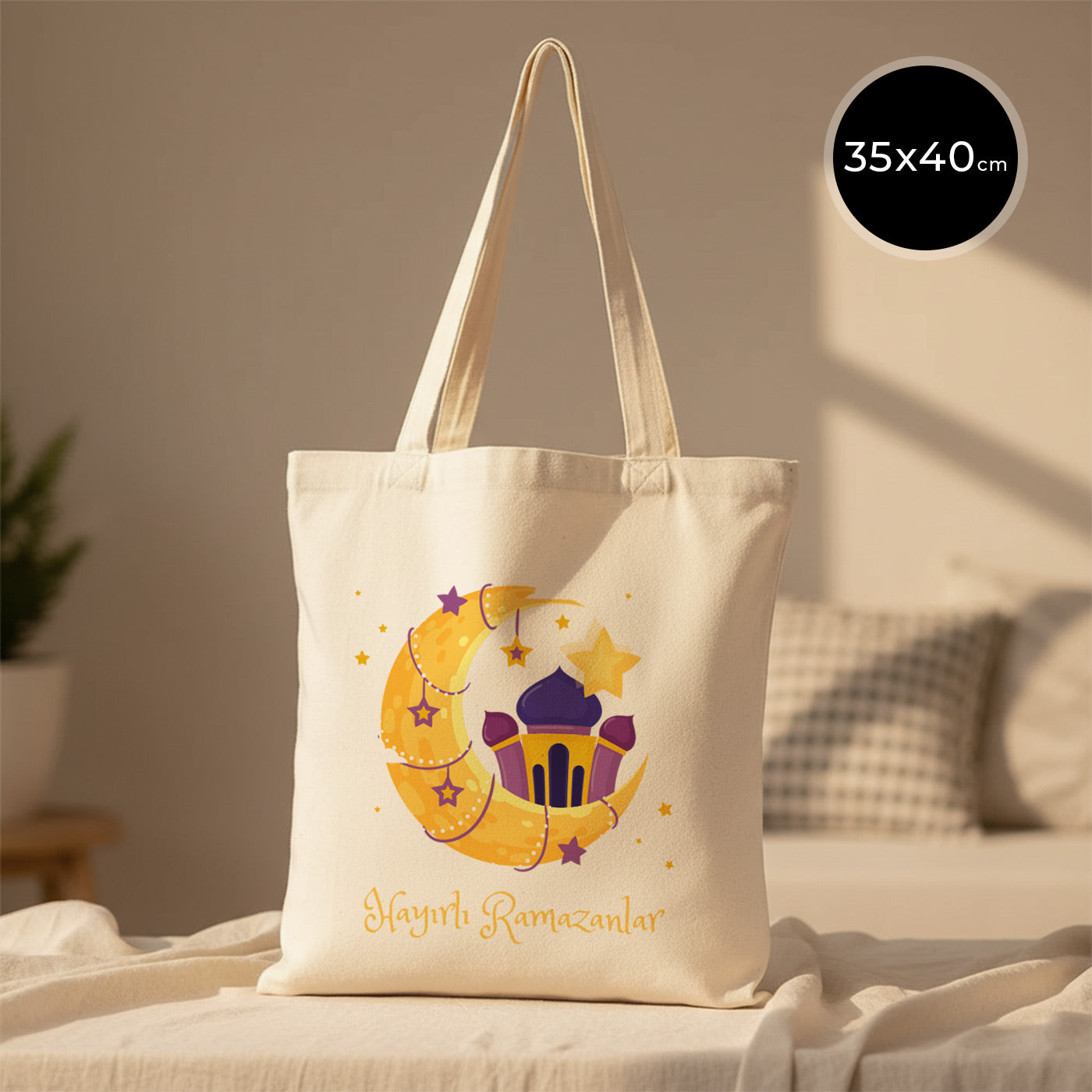 35×40 cm ölçülerinde doğal ham bez tote çanta üzerinde mor fener, sarı hilal ve yıldızlardan oluşan bayram illüstrasyonu ile altında “Hayırlı Ramazanlar” yazısı; arka planda açık renkli kanepe ve desenli yastık.
