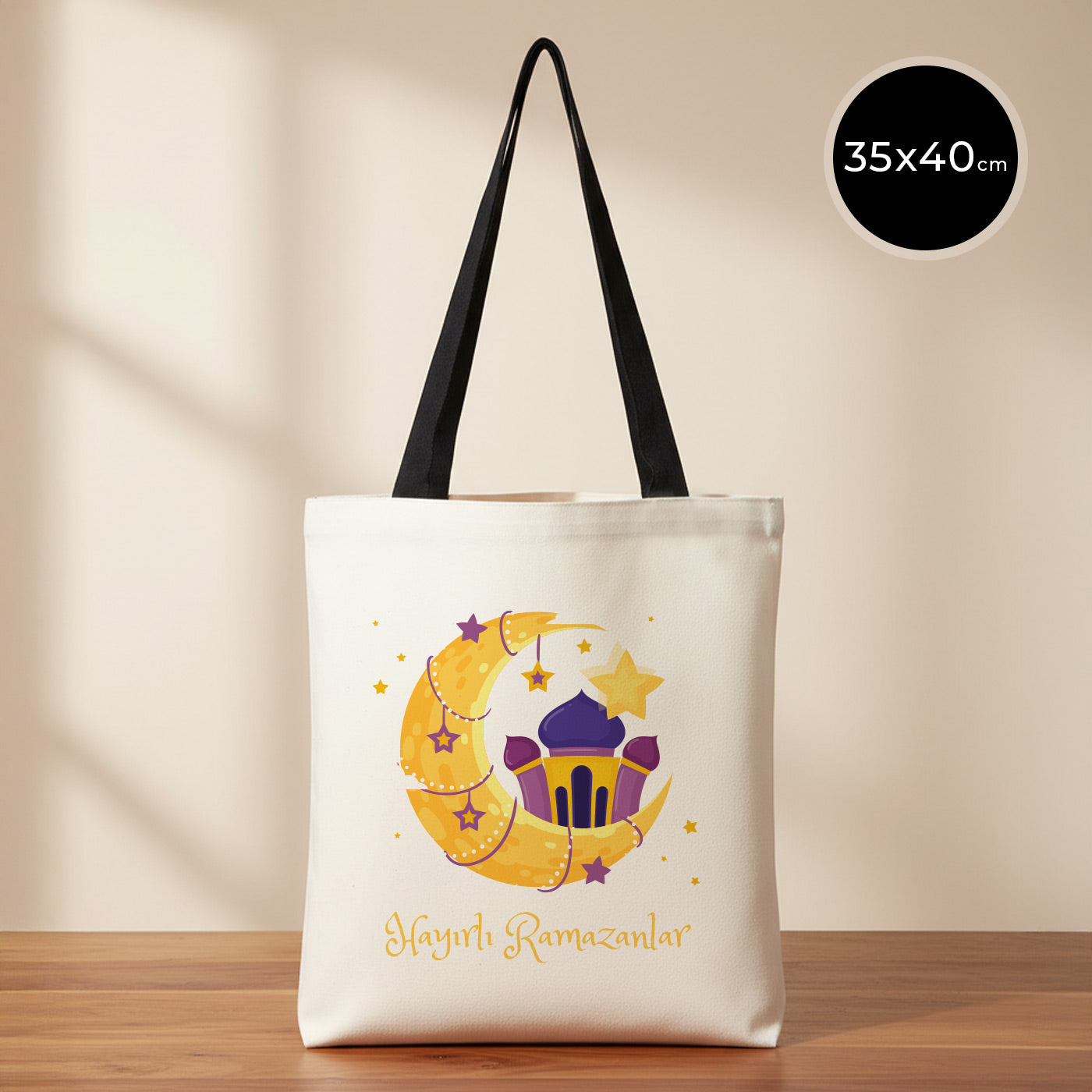 35×40 cm ölçülerinde doğal ham bez tote çanta üzerinde siyah askılar; ön yüzünde mor fener, sarı hilal ve yıldızlardan oluşan bayram illüstrasyonu ile altında “Hayırlı Ramazanlar” yazısı, sade açık renkli arka plan ve ahşap yüzey.