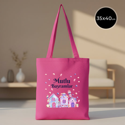 35×40 cm fuşya renk gabardin bez tote çanta üzerinde pastel şeker evleri illüstrasyonu, uçuşan renkli şeker detayları ve üstte koyu mor “Mutlu Bayramlar” yazısı; fuşya omuz askıları ve modern açık tonlu iç mekân arka plan.