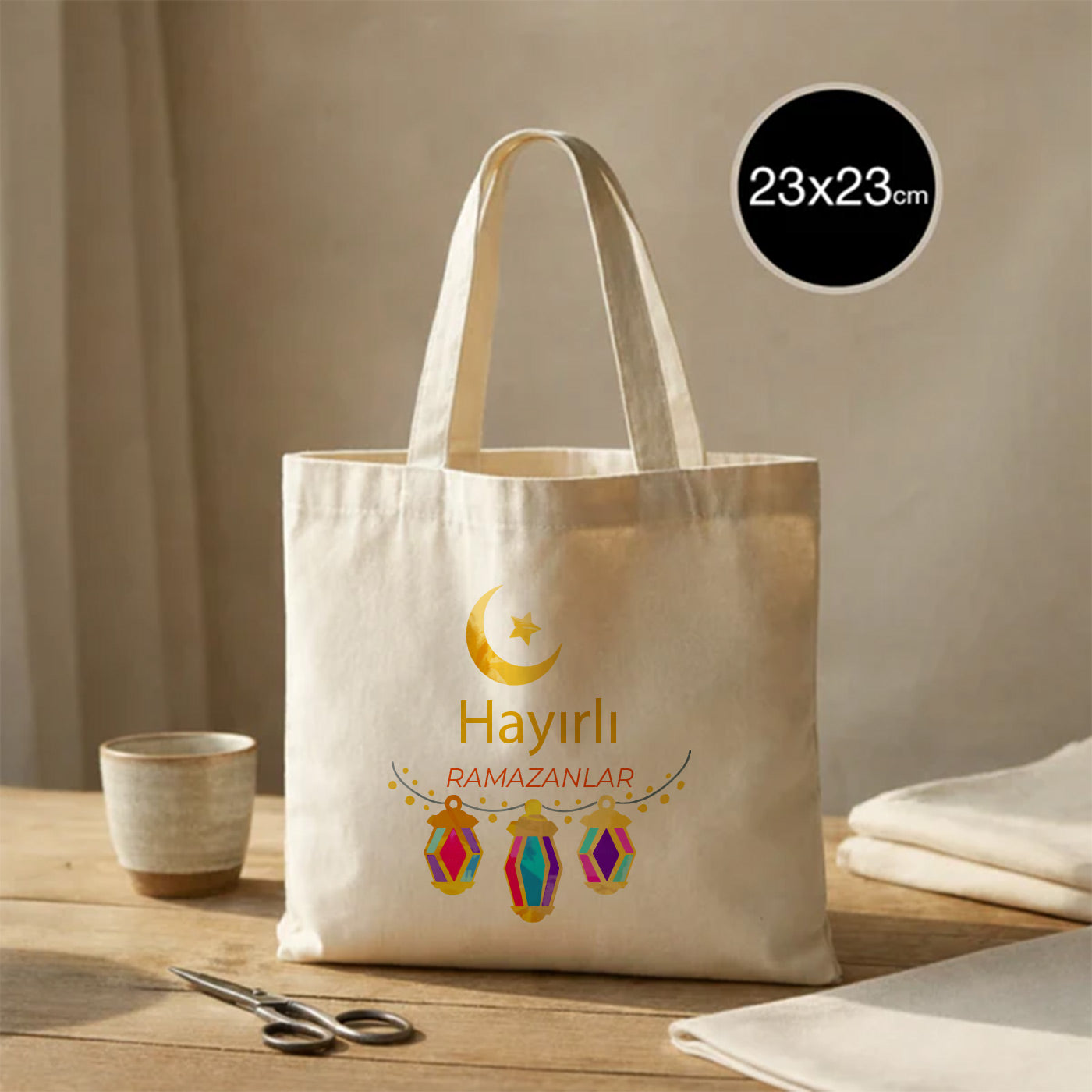 23×23 cm ölçülerinde doğal ham bez mini tote çanta üzerinde üstte sarı hilal ve yıldız, altta üç adet renkli asılı fener illüstrasyonu ve ortada “Hayırlı Ramazanlar” yazısı; ahşap masa üzerinde kahve fincanı, makas ve katlı kumaşların yanında.