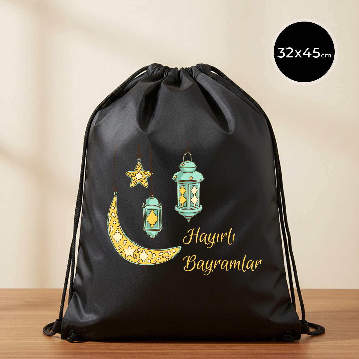 32×45 cm ölçülerinde siyah ip büzgülü bez sırt çantası üzerinde asılı turkuaz fenerler, süslemeli sarı hilal ve yıldız illüstrasyonu ile sağ altta “Hayırlı Bayramlar” yazısı; sade açık renkli arka plan ve ahşap yüzey.