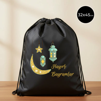 32×45 cm ölçülerinde siyah ip büzgülü bez sırt çantası üzerinde asılı turkuaz fenerler, süslemeli sarı hilal ve yıldız illüstrasyonu ile sağ altta “Hayırlı Bayramlar” yazısı; sade açık renkli arka plan ve ahşap yüzey.